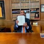 A person sits at a desk in an office holding a signed document related to the Canada Mutual Recognition Agreement. Bookshelves and framed items are visible in the background.