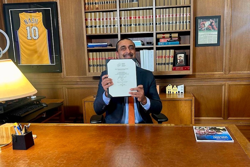 A person sits at a desk in an office holding a signed document related to the Canada Mutual Recognition Agreement. Bookshelves and framed items are visible in the background.