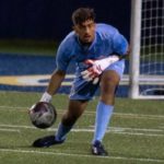 A focused Matheus De Souza, in a light blue keeper kit and gloves, reaches down to collect the ball during a match, with the goal net visible behind him.