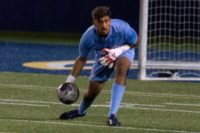 A focused Matheus De Souza, in a light blue keeper kit and gloves, reaches down to collect the ball during a match, with the goal net visible behind him.