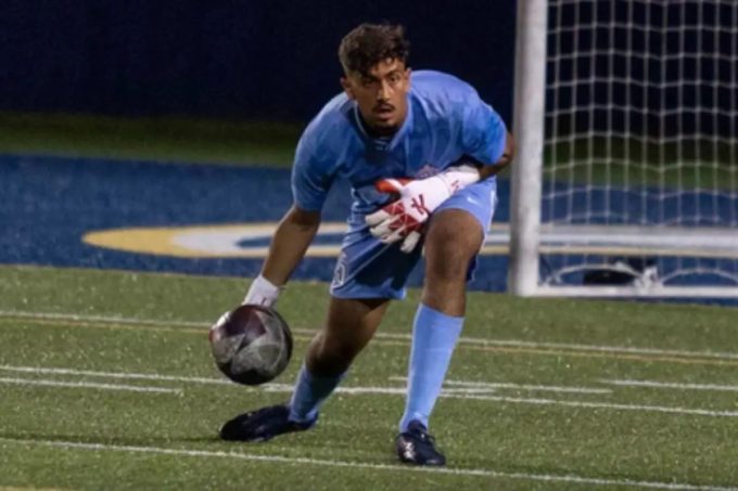 A focused Matheus De Souza, in a light blue keeper kit and gloves, reaches down to collect the ball during a match, with the goal net visible behind him.