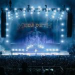 Megadeth performing on a brightly lit concert stage during their farewell tour, with a large crowd watching and the band’s backdrop illuminated in blue lights.