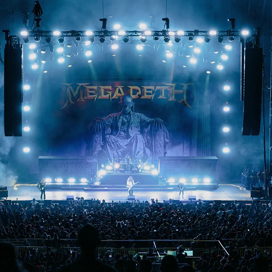 Megadeth performing on a brightly lit concert stage during their farewell tour, with a large crowd watching and the band’s backdrop illuminated in blue lights.