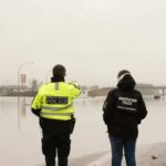 Two Abbotsford police officers stand facing a flooded section of Highway 1, which is completely covered by water on a grey winter day.