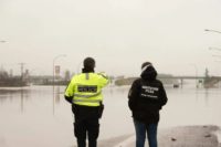 Two Abbotsford police officers stand facing a flooded section of Highway 1, which is completely covered by water on a grey winter day.