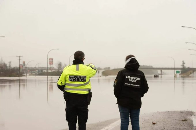 Two Abbotsford police officers stand facing a flooded section of Highway 1, which is completely covered by water on a grey winter day.