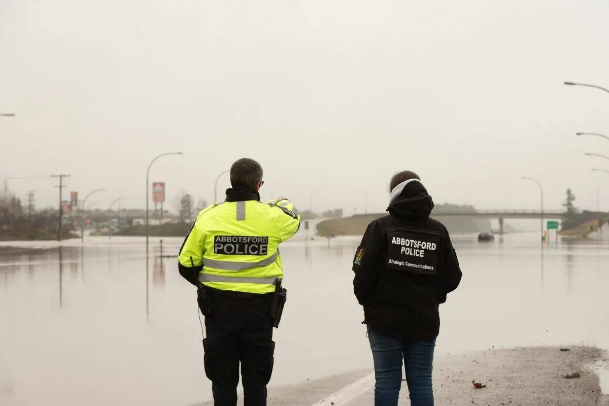 Two Abbotsford police officers stand facing a flooded section of Highway 1, which is completely covered by water on a grey winter day.