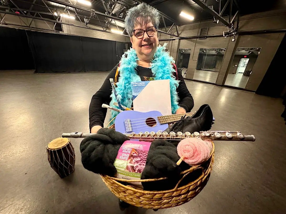 A woman stands in a studio holding a basket of yarn, a flute, a ukulele and other creative supplies that reflect the Artist Garage Sale in Surrey.