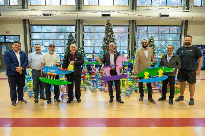 Community partners stand in a gym holding colourful wooden rocking toys, with rows of student-built toys for the Surrey Christmas Bureau behind them.