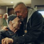 Two men share an emotional embrace in a lounge area, capturing a moment from the B.C. documentary Good to Be Seen about actor Michael Hogan’s recovery.