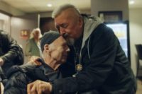 Two men share an emotional embrace in a lounge area, capturing a moment from the B.C. documentary Good to Be Seen about actor Michael Hogan’s recovery.