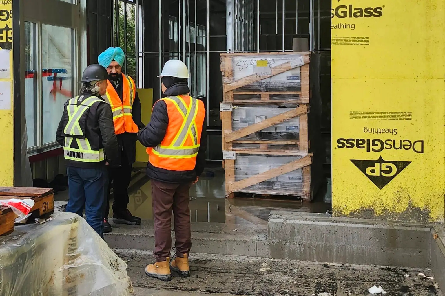 Three people in safety gear stand at a construction entrance discussing progress on the Guru Nanak Diversity Village long-term care facility in Cloverdale.