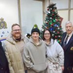 Family members and RCMP veterans stand in front of a Christmas tree and toy donations at the Langley RCMP detachment.
