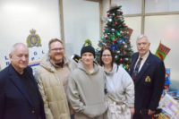 Family members and RCMP veterans stand in front of a Christmas tree and toy donations at the Langley RCMP detachment.