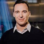 Portrait of teacher Lev Mihalik standing indoors in a dark sweater and collared shirt, winner of UBC’s 2025 Alumni Educator of the Year Award.