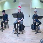 Five actors sit on stools with music stands and microphones in a bright rehearsal room during a Naked Stage readers theatre performance.