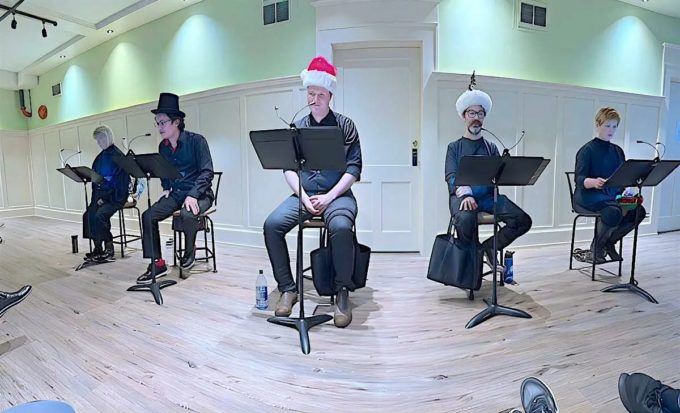 Five actors sit on stools with music stands and microphones in a bright rehearsal room during a Naked Stage readers theatre performance.
