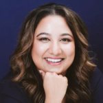 Smiling woman with wavy brown hair posing against a dark blue background, identified as Surrey educator Rochelle D. Prasad.