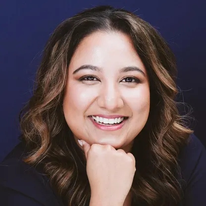Smiling woman with wavy brown hair posing against a dark blue background, identified as Surrey educator Rochelle D. Prasad.