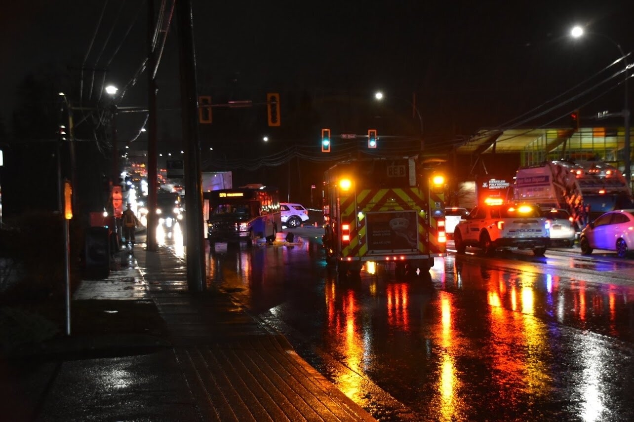 Emergency vehicles and police block a wet Surrey intersection at night after a shuttle bus collision.