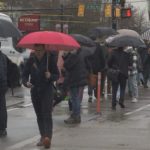 People walk across a busy city intersection under umbrellas during heavy rain, illustrating the incoming storm in Metro Vancouver.