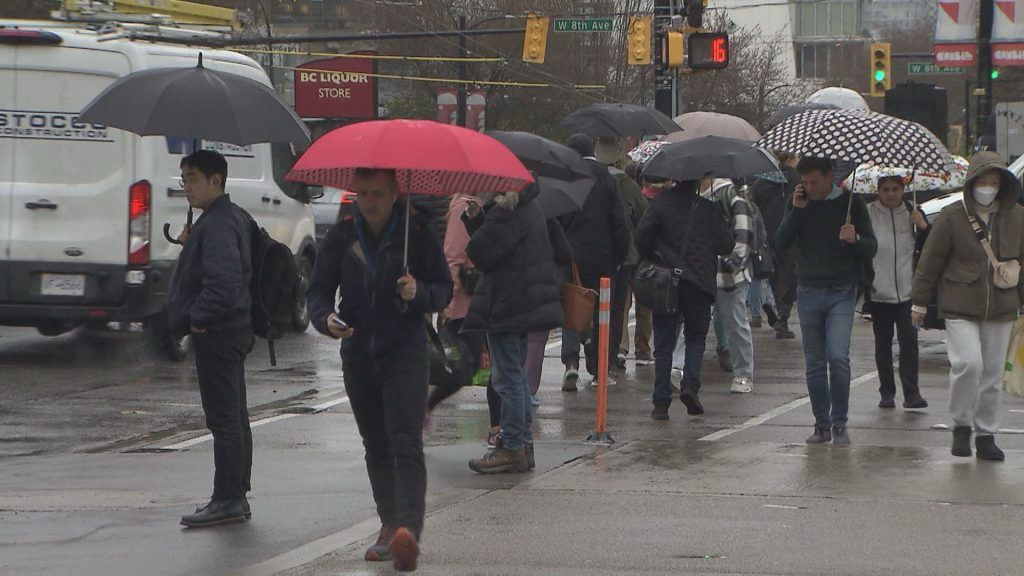 People walk across a busy city intersection under umbrellas during heavy rain, illustrating the incoming storm in Metro Vancouver.