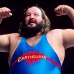 Professional wrestler in a blue “Earthquake” singlet poses with both arms flexed, highlighting his strength in the ring.