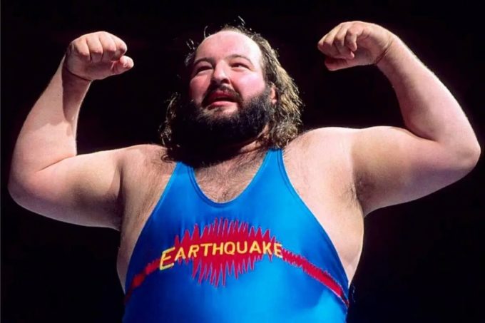 Professional wrestler in a blue “Earthquake” singlet poses with both arms flexed, highlighting his strength in the ring.