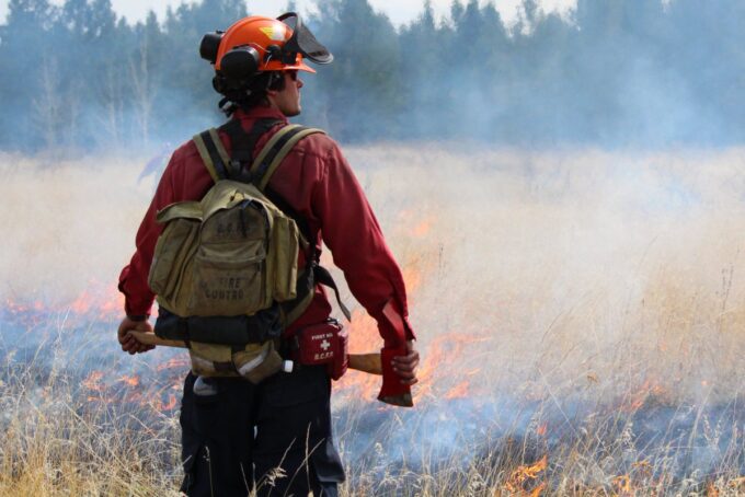 Dark rain clouds moving over a wildfire-affected forest area in British Columbia as smoke rises in the background.